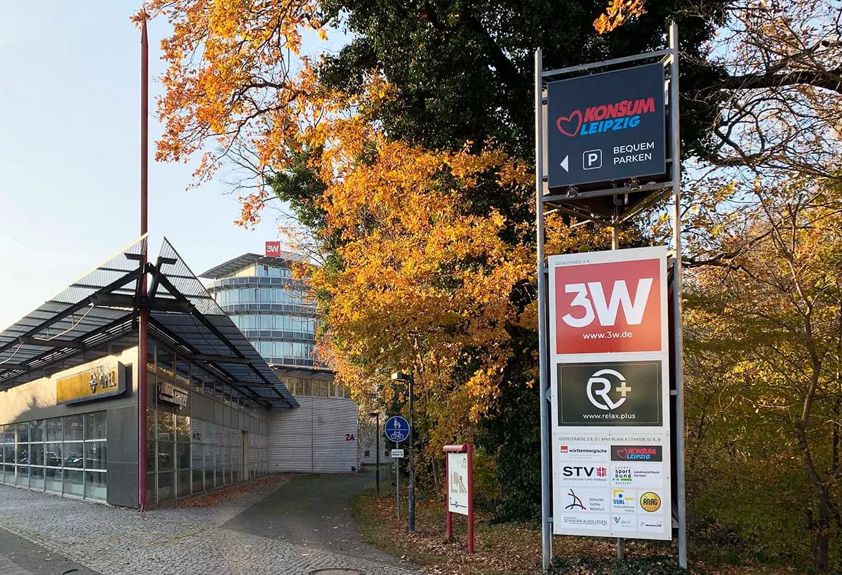 Straßenszene mit einem großen Werbeschild an einem Gehweg, umgeben von herbstlich gefärbten Bäumen. Auf dem Schild sind unter anderem „KONSUM Leipzig“, „3W“ und „R+“ zu lesen. Im Hintergrund befindet sich ein modernes Bürogebäude mit Glasfassade, darüber ein rotes „3W“-Logo.