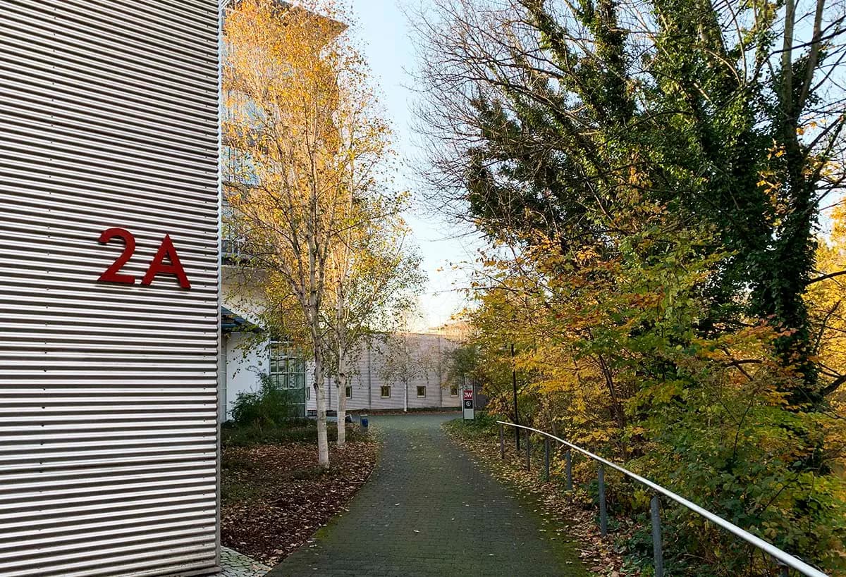 Blick auf einen gepflasterten Fußweg zwischen modernen Gebäuden, gesäumt von herbstlich gefärbten Bäumen mit gelbem Laub. Links ist eine Metallfassade mit roter Aufschrift „2A“ zu sehen, im Hintergrund weitere Gebäude und ein Hinweisschild.
