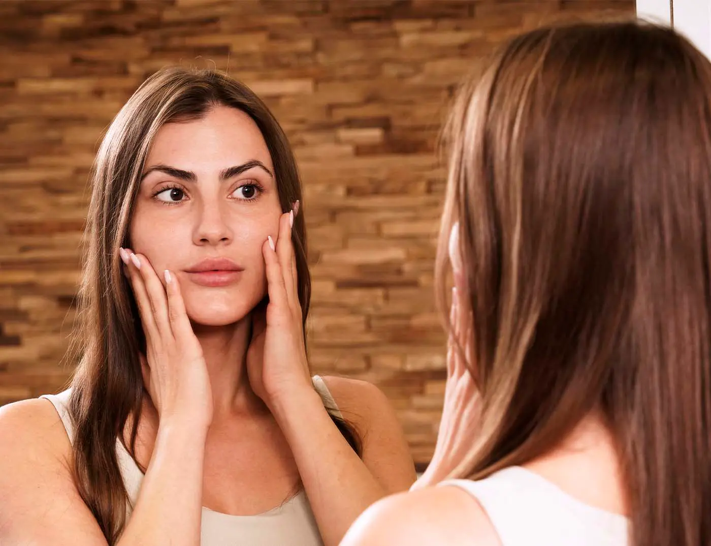 Hübsche, junge Frau betrachtet ihr Gesicht im Spiegel mit den Händen an ihren Wangen