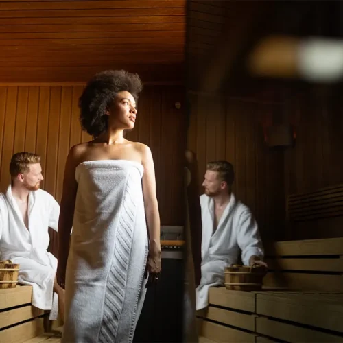 Eine Frau mit Afrofrisur steht in einem Handtuch gewickelt in einer holzvertäfelten Sauna, während zwei Männer in weißen Bademänteln auf den Holzbänken sitzen und Holzeimer neben sich stehen haben.