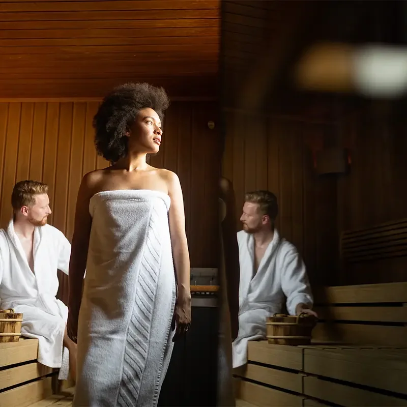 Eine Frau mit Afrofrisur steht in einem Handtuch gewickelt in einer holzvertäfelten Sauna, während zwei Männer in weißen Bademänteln auf den Holzbänken sitzen und Holzeimer neben sich stehen haben.