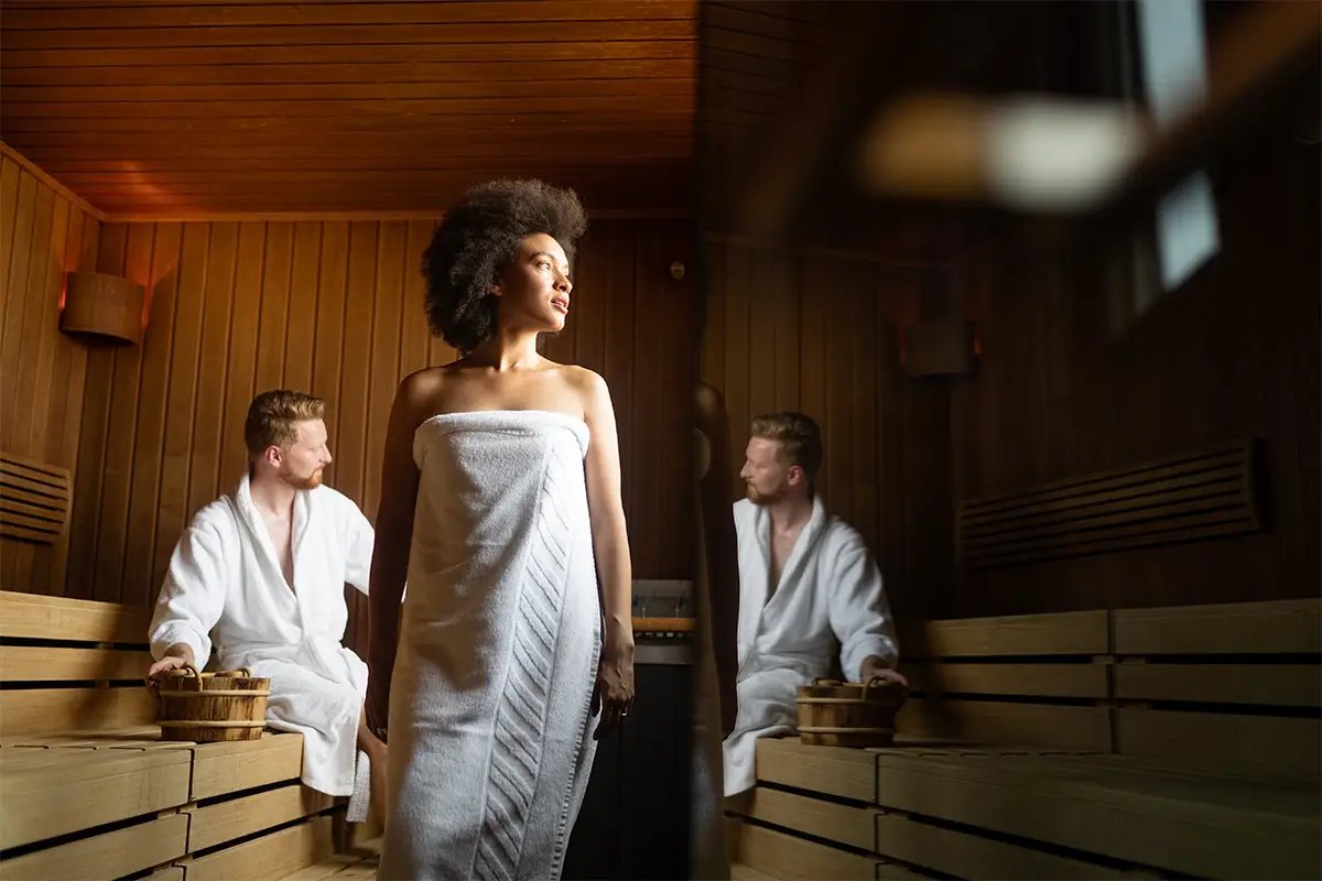 Eine Frau mit Afrofrisur steht in einem Handtuch gewickelt in einer holzvertäfelten Sauna, während zwei Männer in weißen Bademänteln auf den Holzbänken sitzen und Holzeimer neben sich stehen haben.
