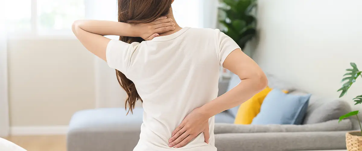 Frau sitzt in einem Wohnzimmer und hält sich Nacken und unteren Rücken vor Schmerzen.