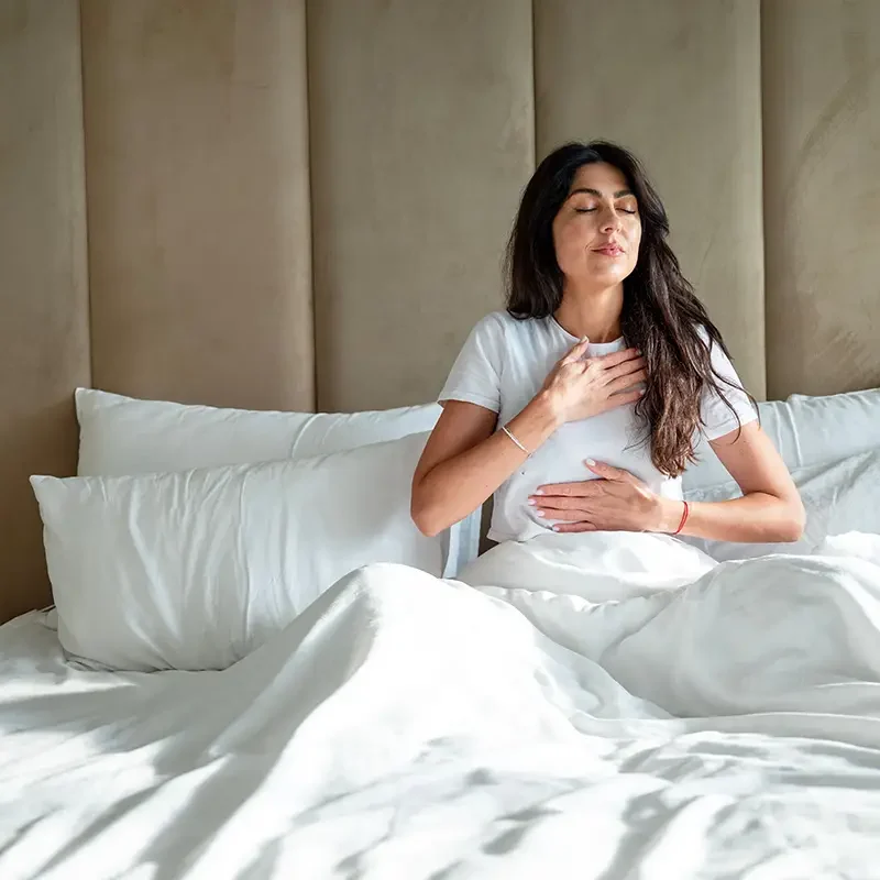 Eine Frau sitzt aufrecht im Bett mit weißer Bettwäsche und geschlossenen Augen, während sie eine Hand auf die Brust und die andere auf den Bauch legt, als würde sie bewusst tief atmen.