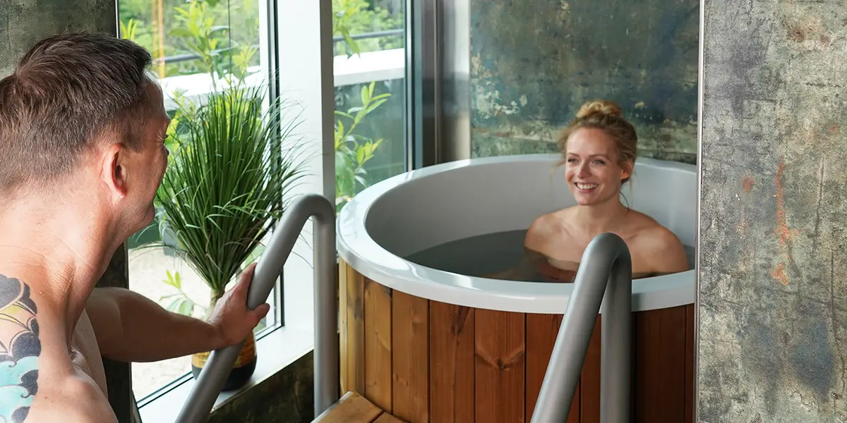 Ein Mann mit Tätowierung spricht mit einer lächelnden Frau, die in einem runden Holz-Tauchbecken sitzt, neben einem Fenster mit Pflanzen.