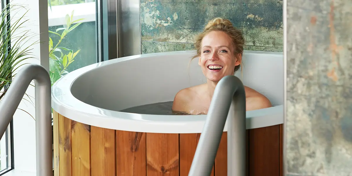 Lachende Frau sitzt in einem runden Holz-Tauchbecken mit warmem Wasser, neben einem Fenster mit Pflanzen.