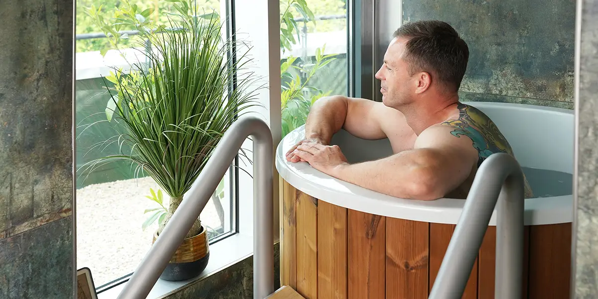 Ein Mann mit Tätowierung entspannt in einem runden Holz-Tauchbecken und blickt aus dem Fenster auf eine grüne Pflanze.