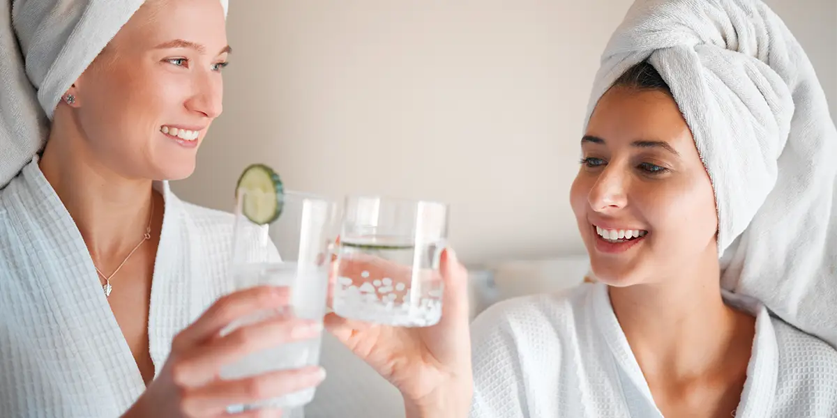 Zwei Frauen im Bademantel mit Turban stoßen nach dem Saunagang mit einem Glas Wasser an und lachen entspannt.